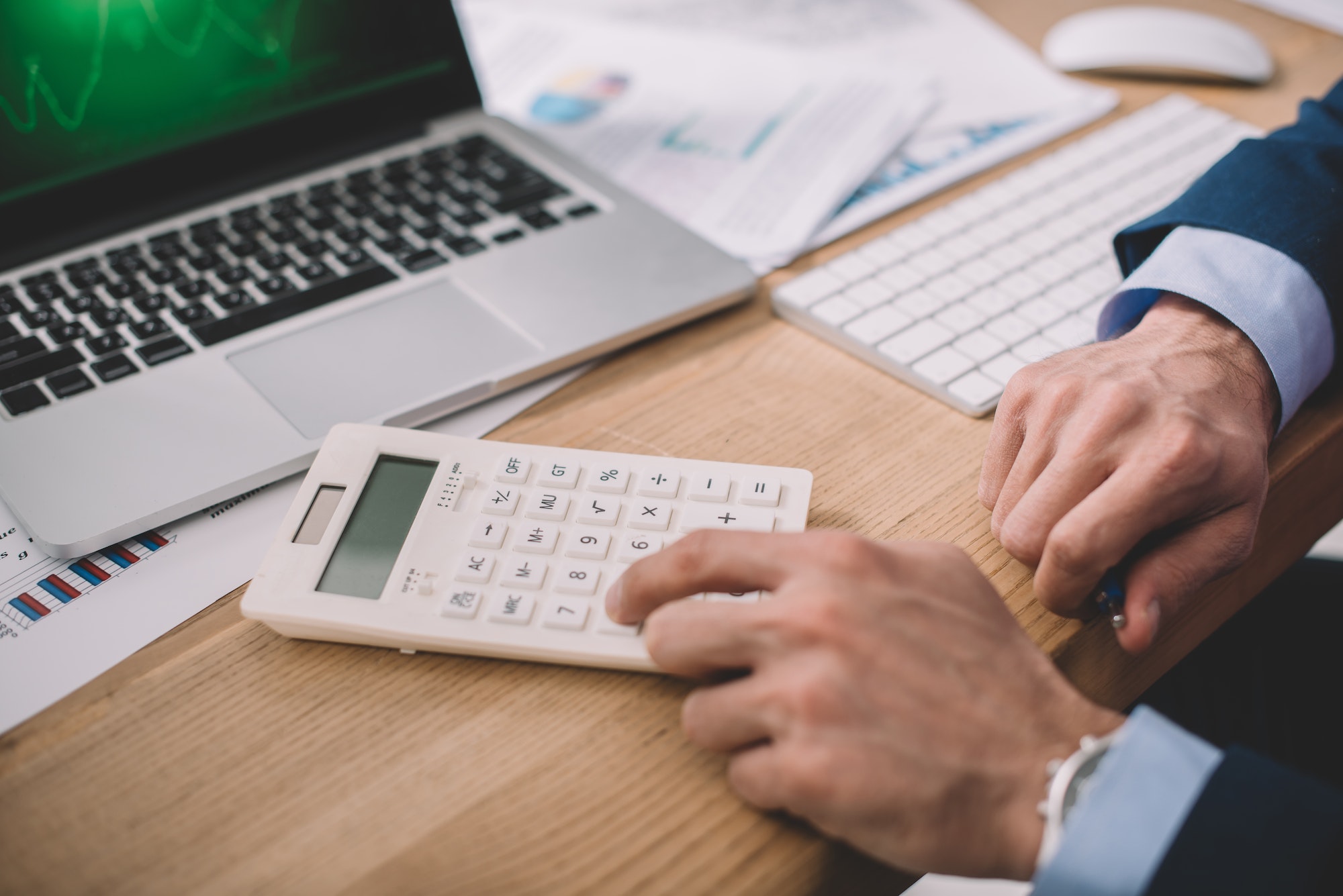 Cropped view of data analyst using calculator near laptop and papers on table